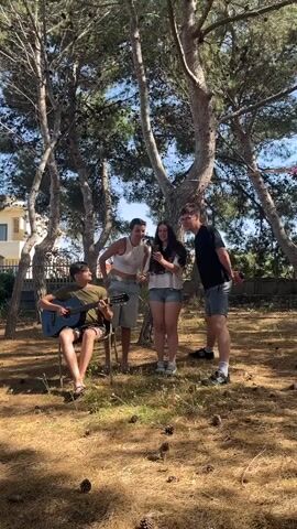 Four people stand outdoors in what appears to be a yard or wooded area. One person is sitting on a metal folding chair, playing an acoustic guitar. Three people stand together, watching the person playing the guitar. The person in the center appears to be holding a smartphone and looking at the screen. The person to the left of the smartphone holder laughs, while the person on the right is also smiling. The ground is covered in dry grass and scattered pine cones. Several tall, thin trees with sparse foliage provide shade. A building is visible in the background, beyond a low fence. The sky is clear and blue. The group seems to be enjoying a casual, musical gathering.