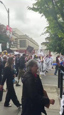 A group of people, some wearing white and black outfits, walk down a street. Many people are holding white flags and some are wearing flags around their necks. One person is wearing yellow boots. Trees are visible on the right side of the frame, and a building is visible on the left. The sky is overcast.
