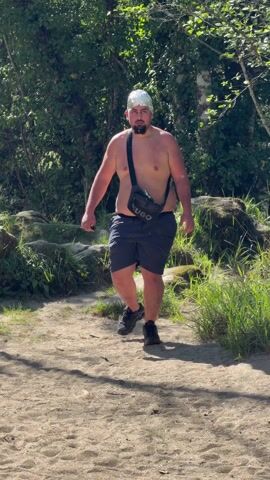A man walks toward the camera in a wooded area. He is shirtless and wearing dark shorts, black sneakers, and a silver foil beanie. He also has a black shoulder bag. The man walks along a dirt path on a sunny day. Behind him is a lush green area with trees, grass, and some rocks. The man appears to be looking at something in his hand, then he looks up and walks toward the camera.