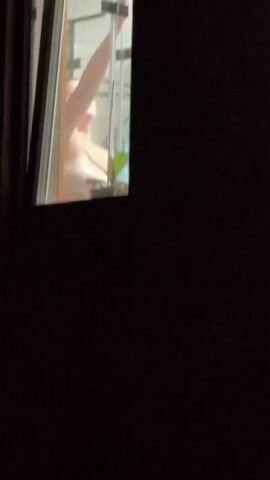 A video captures a person in a bathroom from the exterior of a window at night. The person, who appears to be female, is partially visible through the window, possibly in the process of showering or bathing, as indicated by the presence of a showerhead and what seems to be a shower curtain. A potted plant is also visible on a ledge inside the bathroom. The lighting inside the bathroom appears to be on, illuminating the scene, while the exterior is dark.