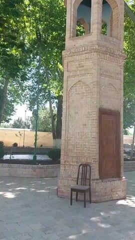 The video shows an outdoor scene with a brick tower with a door and arched openings at the top. A simple chair sits at the base of the tower. Behind the tower is a raised brick-lined pool or trough with water. The area is shaded by large trees with dense green leaves. The camera pans upward to show the full height of the tower and then focuses on the large trees, revealing their multiple trunks emerging from the same base, surrounded by a brick planter. The video is shot in daylight with sunlight filtering through the leaves.