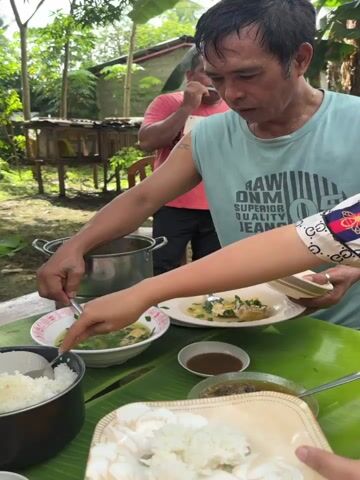 A person wearing a light blue sleeveless shirt is serving food from a large metal pot into a white bowl with a spoon. Another person, whose hands are visible, is also serving food onto a plate, which the person in the light blue shirt is holding. On the table, which is covered with green banana leaves, there are several dishes, including a pot of white rice, two bowls of soup with greens, two plates of meat, and two small bowls of brown sauce. A man in a red t-shirt is standing behind them, holding a plate of food. The scene takes place outdoors, with trees and a house in the background.