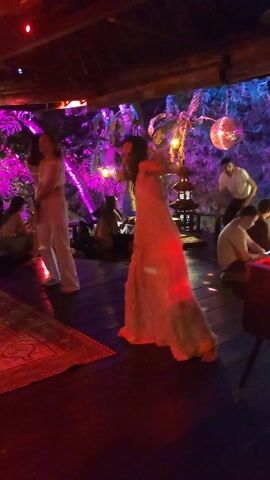 A woman in a long white dress is dancing on a wooden floor, illuminated by purple lighting. Another woman in a white two-piece outfit dances beside her. Several other individuals are seated or standing around the edges of the room, observing or engaging in quiet activities. Overhead, decorative lights and what appears to be foliage create a festive atmosphere. A patterned rug lies on the floor near the dancing women. A man in a white shirt stands in the background with his hands on his hips, watching the dancers. Another man is sitting with a laptop.