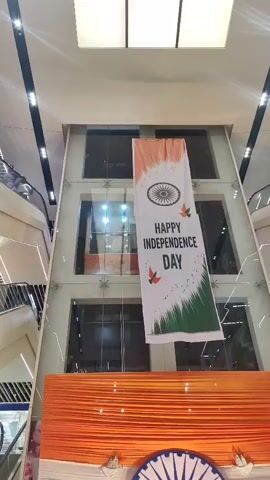 This video displays a large banner hanging in a mall, celebrating Indian Independence Day. The banner features the Indian flag's colors and the text "HAPPY INDEPENDENCE DAY." Below the banner, a decorative display mimicking the Indian flag is set up, with a large Ashoka Chakra. A young man stands in front of the display, posing for a photo. The environment is an indoor shopping mall with escalators visible in the background.