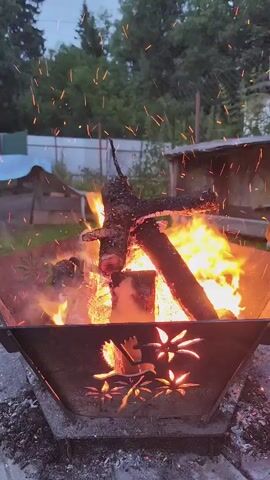 The video shows a close-up of a bonfire in a decorative metal fire pit. The fire pit is octagonal in shape with intricate cutouts of leaves and flowers on its sides. Logs are burning intensely within the pit, with flames reaching upwards and sparks flying into the air. The background is blurred but appears to be an outdoor setting at dusk, with trees and a fence visible. The sky is a dusky blue, indicating that it is either early morning or late evening. The overall mood is warm and inviting, suggesting a cozy evening outdoors. There is no visible text in the video.