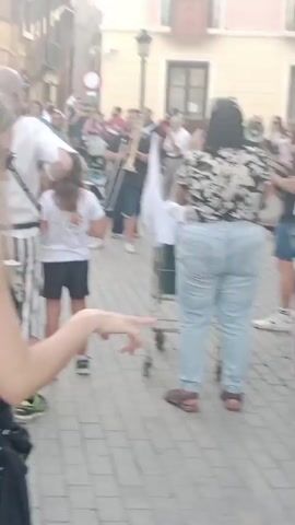 The video shows a street celebration with a group of people. In the foreground, a woman wearing a floral shirt and light blue jeans is pushing a shopping cart. She is facing away from the camera. To her left, a young girl in a white t-shirt and black shorts is standing. Behind the woman and to her left, a band is playing music. One person is playing a trombone, another a drum, and someone else is holding a megaphone. There are other people in the background, some watching the band and others walking. The setting appears to be a town square or street paved with cobblestones, with buildings on either side. The lighting suggests it is daytime. The overall atmosphere seems festive.