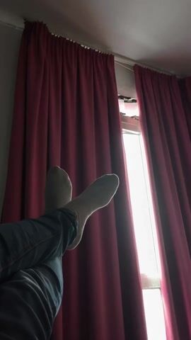 The shot is an upward view of a pair of crossed legs against a window with dark red curtains. The person is wearing light-colored socks and blue jeans. The window is allowing a bright, diffused light to enter the room. The focus is primarily on the socks and feet. The environment appears to be indoors.