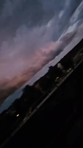 A video shows a tilted view of a sky filled with dark, ominous clouds tinged with purple and pink hues. The clouds are dense and appear to be moving, suggesting a storm. Below the sky, a silhouetted landscape of trees and a faint outline of buildings is visible. The bottom of the frame is dominated by a dark, blurry foreground, possibly a fence or railing, with a faint circular object visible in the lower left quadrant. The lighting suggests it is dusk or twilight.