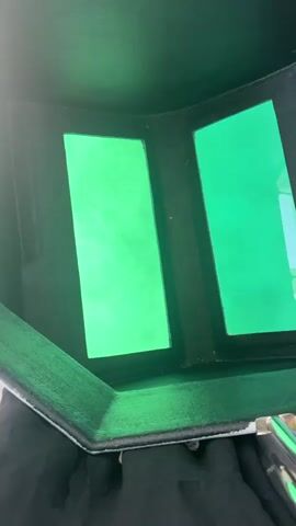 The video shows a close-up, interior view of a welding helmet. The camera is looking through the two green tinted windows of the helmet. The interior of the helmet appears to be black and green. Some faint light or dust can be seen on the left window. The helmet is angled downwards, suggesting it is being held or worn. The overall impression is a very limited view through the protective lenses of the welding mask.