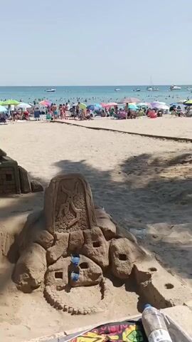 The video pans across a beach with a large, intricate sandcastle in the foreground. The sandcastle is multi-tiered and features detailed architectural elements such as stairs, walls, and turrets. Some of the sandcastle structures resemble ancient buildings or fortresses. In front of the main sandcastle, there is a smaller, cruder sand sculpture, possibly a base or an earlier attempt. A pair of sunglasses rests on this smaller sculpture. Further back, the beach is populated with numerous people relaxing under colorful umbrellas and sunshades. The ocean is visible in the background with several boats scattered across its surface. The sky is clear and blue, indicating a sunny day. The overall scene depicts a lively beach environment with a focus on the impressive sandcastle art.