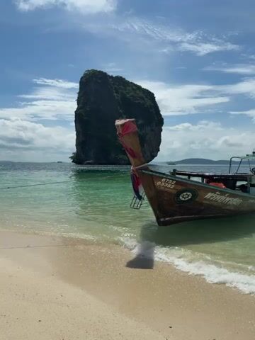 The video opens with a shot of a long-tail boat moored in shallow, clear turquoise water near a sandy beach. A large, jungle-covered rock formation rises dramatically from the sea in the background. The boat is brown with some blue trim and has Thai writing on its side. The sun is shining, and there are white clouds scattered across a bright blue sky. As the video progresses, the camera pans to the right, showing more of the water and the beach. Another long-tail boat is visible in the distance on the right side of the frame. The waves gently lap at the shore. The overall scene is one of a serene tropical paradise.