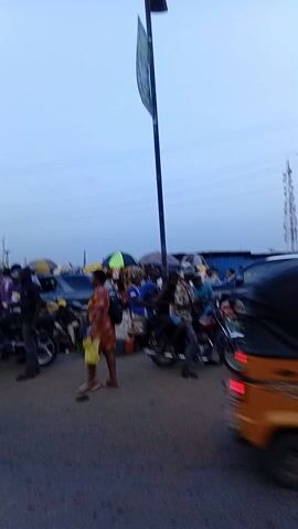 The video shows a busy street scene with people, vehicles, and street vendors. There are cars, motorcycles, and auto-rickshaws moving through the area. People are walking around, some appearing to be shopping. Street vendors have set up stands with goods for sale, including clothing and bags. A sign warns against parking, waiting, or stopping in the area. The sky is overcast and the lighting suggests it might be early morning or late afternoon. The environment appears to be urban, with buildings visible in the background and power lines overhead.