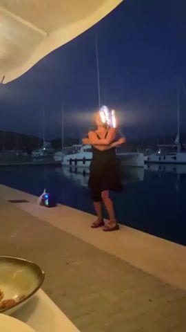 A woman wearing a black dress and a red hat is performing with fire. She is holding two flaming objects, one in each hand, and appears to be swinging them in a circular motion. The performance is taking place outdoors at night, with a body of water and several sailboats visible in the background. There is also a small blue speaker on the ground near the woman.