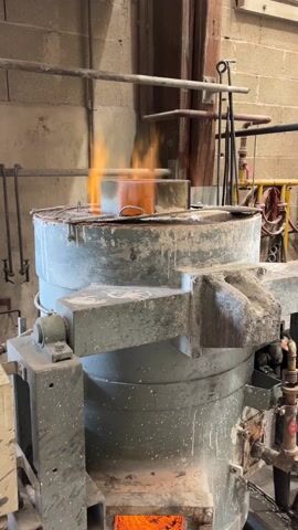 The video captures a large, grey industrial furnace in full operation within what appears to be a foundry or workshop. From the top opening of the cylindrical furnace, vivid orange flames erupt, indicating the presence of intense heat. A lower, rectangular opening on the side of the furnace reveals a bright, glowing orange-red interior, consistent with molten material or a superheated chamber.

The furnace itself is robust, showing signs of wear and flecked with white paint splatters. In the background, brick walls, metal pipes, and various industrial fixtures are visible, reinforcing the setting as an industrial facility. The floor surrounding the base of the furnace is covered with debris. The scene is accompanied by an audible hum and roar, indicative of intense heat and operational machinery. There is no visible text in the video.

This industrial process is taking place in Saint-Rémy-lès-Chevreuse, France.
