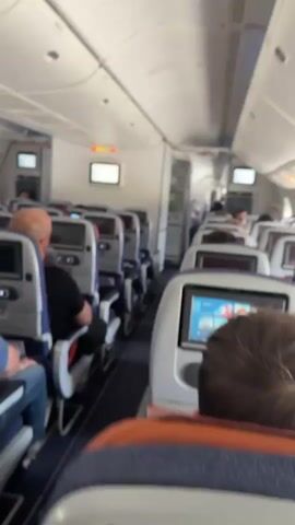 The video pans through the interior of an airplane cabin. Rows of passenger seats are visible, most equipped with personal entertainment screens. Several passengers are seated, facing forward or looking at their screens. The overhead compartments and cabin lighting are also visible. The lighting suggests it is daytime, with sunlight entering through the windows. The aisle is carpeted in dark blue. Some screens display colorful content. The camera movement is steady at times and slightly jerky when moving to a new angle. The overall impression is of a typical commercial flight cabin before takeoff or during a quiet phase of the flight.