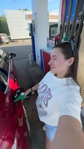 A young woman is taking a selfie while refueling a red car at a gas station. She's wearing a white t-shirt, denim shorts, and white sneakers. Sunglasses are perched on her head. The gas pump nozzle is inserted into the car's fuel tank. The gas station has a modern design, with other vehicles parked nearby. The sky is clear and the lighting is bright, suggesting it's daytime.