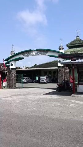 The video begins with a view of an arched gateway with an inscription and minarets on either side. To the right, a mosque with a green tiled roof is visible. In front of the gateway, there is a paved area with cars and a motorcycle parked. A person rides a red motorcycle from left to right across the frame. Further to the left, a covered stall or market area is visible with several people and motorcycles. The camera pans to the right, showing more of the stall area, which appears to be selling goods, with signs advertising various items. The sky is blue with some clouds. The video captures a street scene with a mosque and a commercial area.
