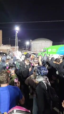 The video depicts a large gathering of people outdoors at night, illuminated by streetlights and other artificial sources. The crowd is dense, with many individuals wearing dark clothing, including hoodies and jackets. A significant number of participants are equipped with protective gear such as gas masks or respirators, and some are wearing helmets. Several people are holding up smartphones, seemingly recording the event.

In the foreground, a person in a blue t-shirt and another with a black bandana covering their face are visible. Backpacks are common among the attendees. In the background, industrial-style brick buildings with a prominent tall brick chimney are discernible. Overhead power lines, utility poles, and streetlights are visible against the dark sky. To the right, a large green and blue truck or tanker, partially visible, displays the text "SUMMA ENERGY." A white vehicle is also seen behind the crowd. On the far left, a uniformed police officer is present, facing the assembled group.

The presence of protective gear suggests a potentially tense atmosphere or preparation for possible confrontations. Sounds of chanting or shouting are audible throughout the video, indicating an active protest or demonstration. The setting appears to be an urban or industrial area at night.