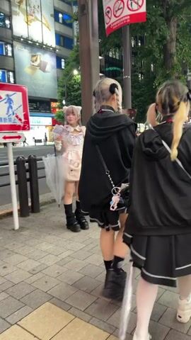 At 00:00, a young woman with blonde hair styled in pigtails, wearing a light pink dress with ruffled trim, cat ears, and knee-high black socks with platform boots, stands on a sidewalk. She holds a clear umbrella and appears to be looking at something off-camera. Behind her, another young woman, also with dark hair, wears a similar pink dress and black platform boots, and holds a book or pamphlet in front of her face. To the right, a third person, with blonde hair and black clothing including a hoodie and skirt, walks away from the camera, also wearing platform boots. The environment is an urban street at night, with buildings, streetlights, and signage visible in the background. A bicycle is parked nearby. At 00:01, the first two women are still in the same positions. The first woman is now holding her umbrella lower and a pamphlet is visible in front of her face. The second woman continues to hold a pamphlet in front of her face. The third person is further away and walking away. At 00:02, the scene remains largely unchanged, with the first two women continuing to hold pamphlets. The bicycle is still visible. At 00:03, the scene shows a wider view of the street. The first two women are less prominent. The third person is now moving further away.