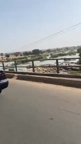 The video shows a dark blue Toyota Camry driving across a bridge. The river below is wide and shallow, with some vegetation and sandbars visible. The sky is clear and pale blue, and the landscape in the background appears dry and sparsely vegetated. The car's license plate is partially visible. The video is shot from the passenger side of the car.