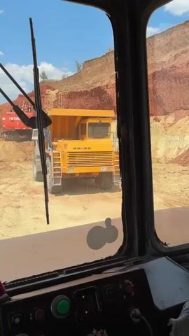 The video provides a view from inside the cabin of a vehicle, looking out through the windshield. A large, yellow dump truck is prominently positioned in the foreground, facing away from the camera. Its rear end is visible, with the number '41' painted on its side. Behind the dump truck, a red excavator is partially visible, positioned on a slope. The environment appears to be a mine or quarry, with large piles of red earth and a clear blue sky overhead. The camera is stationary, capturing the scene with minimal movement.