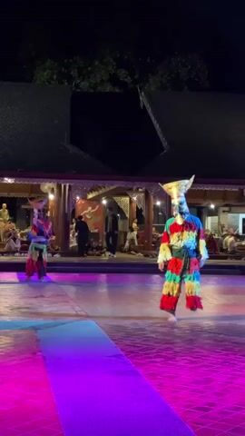 Two performers in colorful, traditional costumes and masks are dancing on a paved outdoor stage at night. The stage is lit with vibrant colored lights. In the background, there is a building with a dark roof and open sides, and an audience is seated. The performers' costumes are made of brightly colored fabric strips and they wear tall, elaborately decorated masks. The performer on the right is facing the camera and appears to be moving their arms and legs in a rhythmic dance. The performer on the left is facing away from the camera and is also dancing. Trees are visible in the dark background above the building.