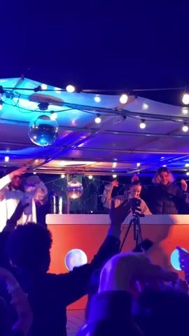 The video shows a group of people gathered in what appears to be an open-air event, possibly a party or concert. The crowd is in the foreground, facing towards a raised platform where several individuals are positioned. On the platform, there’s a DJ booth, behind which at least three people are standing. One person seems to be operating the DJ equipment, while others are interacting with the crowd, waving and gesturing. There is a camera on a tripod, suggesting that the event may be being recorded or live-streamed. The area is decorated with string lights and disco balls hanging from the ceiling, which contribute to the festive atmosphere. The lighting is predominantly blue and purple, adding to the ambiance. The crowd is lively, with people holding up their phones, likely to take pictures or videos. The overall scene conveys a sense of celebration and engagement.