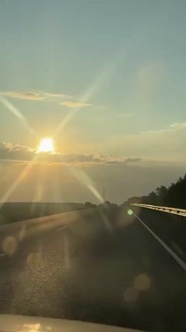 The video is a first-person perspective shot from inside a vehicle traveling on a highway. The sun is setting, casting a warm, golden light across the scene. The sun is partially obscured by clouds and is creating lens flares that streak across the sky and the road. The highway has two lanes of travel and is bordered by a metal guardrail on the right side. To the left of the highway, there is a flat, open field. On the right side, there is a line of trees. The sky is a clear blue with scattered white clouds. The road surface appears to be asphalt and is wet in places, reflecting the sunlight. There are no other vehicles visible on the road in the immediate vicinity.