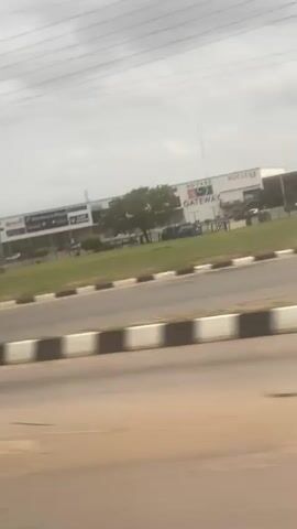 The camera captures a view from a moving vehicle, likely a car, on a road with a grassy median strip. The scene features several buildings set back from the road, including a large shopping center with visible signage for "Shoprite" and "Nucleus". The sky is overcast with grey clouds, and there are numerous power lines running overhead. No people are visible in the shot. The camera is moving at a speed that blurs the foreground slightly, but the buildings in the background are relatively clear. The overall impression is of a commercial area on a cloudy day.