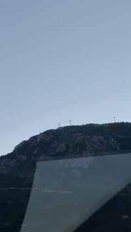 The video shows a dark, rocky mountain against a light blue sky. There are several power line towers visible on the ridge of the mountain. The view is from inside a vehicle, with part of the windshield visible in the lower portion of the frame. The overall lighting suggests it is either early morning or late evening.