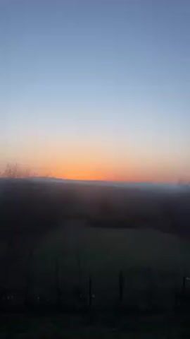 The video shows a landscape at dawn or dusk. The sky transitions from pale blue at the top to orange near the horizon, indicating the presence of the sun. A range of hills or mountains is visible on the horizon. In the foreground, there are silhouettes of trees and foliage, obscuring a small field or open area. A simple fence is also visible at the bottom of the frame.