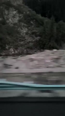 This video shows a blurry landscape from a moving vehicle. The scene appears to be a mountainside with trees and a body of water. The lighting suggests it is either dusk or dawn.