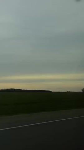 The video displays a static shot from inside a moving vehicle, looking out over a landscape. The primary subjects are a vast, flat, dark green field, likely agricultural, extending from the foreground to the horizon. In the distance, a dark line of trees separates the field from the sky. A two-lane asphalt road with a white dashed dividing line occupies the bottom portion of the frame, indicating the perspective from a moving vehicle. The sky is entirely overcast with heavy gray clouds, although a band of lighter, yellowish-white clouds is visible closer to the horizon, suggesting either early morning or late afternoon. Very faint, small, reddish-pink dots can be seen scattered in the sky, which may be reflections on the glass or sensor artifacts. No people are visible. No text is visible in the frame, but Russian speech is audible in the background. The location is contextualized as Pervomayskiy, Russia.
