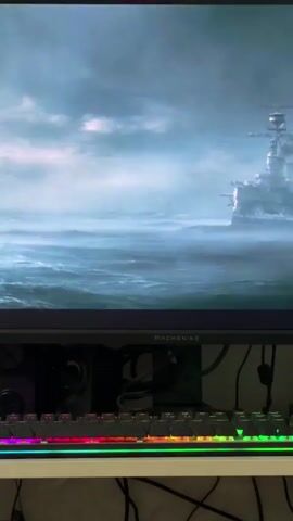 A computer monitor displays an image of a battleship sailing on choppy, gray water under a cloudy sky. Smoke billows from the ship's smokestacks. In the foreground, a computer keyboard with illuminated RGB keys is visible. The scene is static, indicating a paused or still image on the screen.