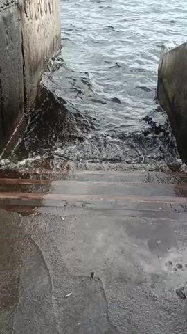 The video shows a set of concrete stairs descending into dark, choppy water. The water is dark and appears to be flowing or being disturbed, creating white foam and splashes as it interacts with the steps. Two concrete walls flank the stairs, forming a narrow channel leading to the water. The overall impression is of a functional, possibly industrial or dockside, entryway to the water, with the water itself appearing somewhat rough or turbulent.
