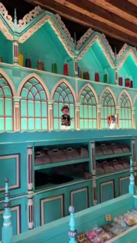 The video depicts the interior of a candy store with a unique, vibrant teal and cream color scheme. The store features elaborate, custom-built shelving units that resemble miniature gothic cathedrals, complete with arched windows and decorative trim. These shelves are stocked with a variety of candies, presented in jars and wrapped packages.

A female employee wearing a white uniform and cap stands behind a counter, presumably assisting customers. Another female customer, dressed in a white top and black pants, is seen browsing the candy selection at one of the shelves. The camera pans around the store, showcasing the detailed interior design, including a wooden beamed ceiling and patterned tile floor.