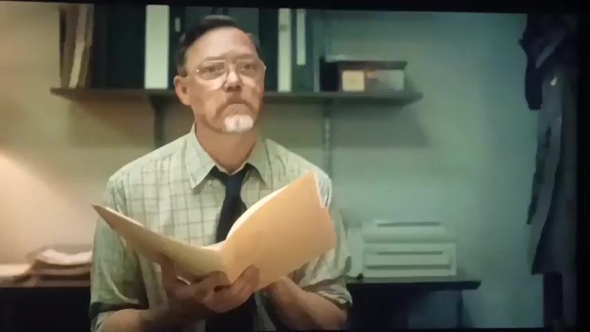 The video begins with a medium shot of a middle-aged white man with short, dark hair, glasses, and a goatee. He is wearing a collared shirt and a tie. He is holding a manila folder open in his hands and looking up and to his left with a serious expression. The background appears to be an office with shelves holding binders and boxes. The camera then cuts to a medium shot of a younger white man with dark, curly hair. He is wearing a collared shirt and a tie. He is looking directly at the camera with a somber expression. The background appears to be an office with blinds on a window and a desk with various items on it. The video continues with slightly different angles of the younger man speaking, with the same somber expression.