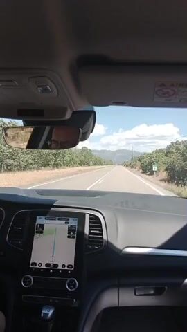 The video is filmed from the perspective of someone in the passenger seat of a car, looking forward through the windshield. The car is moving along a paved road with white lane markings. On either side of the road are trees, likely part of an orchard, with some barren ground visible. In the distance, there are mountains. A GPS navigation system is displayed on a screen in the center of the dashboard. The driver is partially visible in the rearview mirror, wearing glasses. The sun is shining, and there are clouds in the sky. A speed limit sign is visible on the right side of the road.