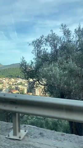 The video shows a view from a moving vehicle looking out over a coastal town. In the foreground, a metal guardrail is visible. Beyond the guardrail, there are buildings with red-tiled roofs and some taller, modern buildings. Green hills covered in trees rise behind the town. The sea is to the right, with a small island visible in the distance. The sky is blue with some white clouds. The sun appears to be shining.