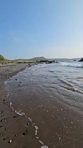 This is a vertical, wide shot of a beach with gentle waves washing ashore on a sunny day. The foreground is a dark, wet sand beach littered with small pebbles and foamy water. Beyond the sand, the ocean stretches out, its surface rippling and reflecting the bright sun. Waves are seen breaking and rolling towards the shore. In the distance, a rocky shoreline with some vegetation can be seen, and the sky above is a clear, pale blue. The sun is high in the sky, casting a bright glare on the water. There are no people or animals visible in the frame.