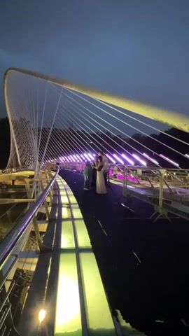The video shows a pedestrian bridge at dusk or night, illuminated by artificial lights. The bridge has a modern design with a large curved structure and numerous cables. The walkway is made of dark material, and sections of the bridge are lit with purple and yellow lights. In the middle of the bridge, three people are visible. One person in a light-colored dress is standing with two other individuals. The person in the dress appears to be posing or being photographed. The background shows trees and possibly buildings in the distance, with a dusky, twilight sky. The lighting suggests a festive or event atmosphere.