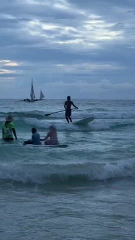 A person on a paddleboard is in the ocean. A sailboat is visible in the distance. Three other people are on paddleboards closer to shore. The sky is cloudy. Waves are breaking on the shore.