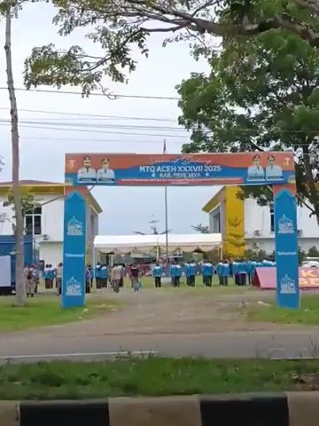 The video opens with a wide shot of an entrance archway decorated with banners. A banner at the top reads "Selamat Datang MTQ ACEH XXXVII 2025 KAB. PIDIE JAYA" with images of two men in suits. Below this, smaller text reads "Mari Kita Tingkatkan SDM Qur'ani Yang Unggul Menuju Aceh Islami Penuh". On the sides of the archway are blue banners with a logo. People are seen standing in a line in front of the archway, some wearing light blue shirts with white collars and white lettering. Others are walking towards a white tent structure in the background. A motorbike is parked on the left side of the frame. The ground is covered with gravel and some grass. The overall scene appears to be an outdoor event or ceremony.