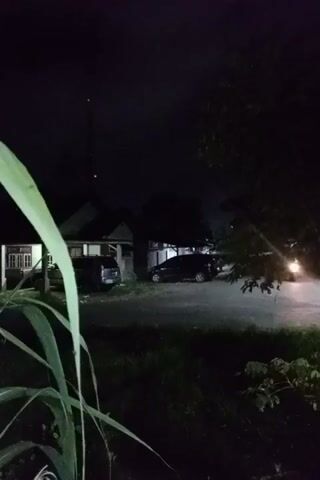 The video shows a still, dark, outdoor scene at night. Two cars are parked in front of houses. Tall grass blades are visible in the foreground to the left. There's a tall antenna in the background, extending upwards from one of the houses. A tree is partially visible on the right. The ambient light illuminates the road and the houses enough to make out basic shapes. The scene seems quiet and still.