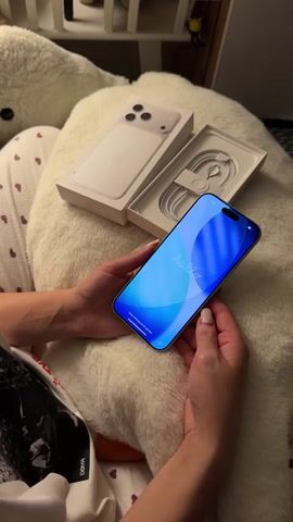 A person is shown holding a new iPhone, which is in its initial setup phase with the word "Halla" displayed on the screen in multiple languages, including Arabic script "مرحبا." The phone appears to be a recent model, possibly an iPhone 15 Pro Max, given its design and the included USB-C charging cable. The device is light-colored, possibly white or natural titanium.

The open packaging for the iPhone is visible next to the person's hands. One half of the box displays the back of the phone, showing the triple camera array and the Apple logo. The other half contains the white charging cable, neatly coiled.

The person is wearing pajamas or loungewear with a white background and small red heart patterns. Part of a word, "DORIA," is visible on their clothing. They are resting their hands on a large, fluffy white pillow or blanket. In the background, a white shelf or piece of furniture can be seen. The lighting suggests an indoor setting, likely a home. The activity depicted is the unboxing and initial setup of a new iPhone.