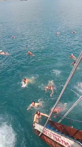 A group of people are swimming in the ocean near a boat. The boat is partially visible in the foreground, with ropes and rigging. Several people are in the water, swimming and splashing. Some are closer to the boat, while others are further away. The water is blue and appears to be clear. In the background, a town or city is visible on the coastline. The sky is bright and sunny.