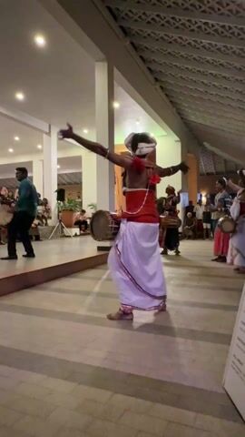 A man in traditional Sri Lankan attire stands in the foreground playing a double-sided drum. He wears a white wrap with red stripes around his waist and a red top, along with a white head covering and red flower accents. He is barefoot and facing away from the camera. To his left, another man in a dark shirt and pants also plays a drum while standing on a raised platform. Several other people, some also with drums, are visible further back. A crowd of seated and standing people watches the performance. The scene is set in a large, open, well-lit space with white pillars supporting a roof, suggesting an open-air event hall or resort area.