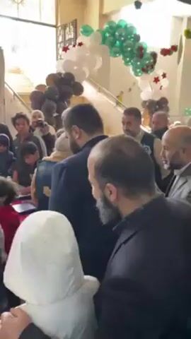 A crowd of people, mostly men and children, are gathered near a decorated staircase. The staircase has a railing and is adorned with a balloon arch of white, green, black, and red star-shaped balloons. Some people are wearing dark suits, while others wear casual clothing and headscarves. One person holds a camera and appears to be taking a photo. A table covered in what looks to be boxes of treats can be seen near the center of the frame. The room is well-lit with natural light coming in from an entranceway or large window.