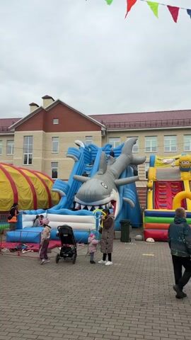 The video shows a festive outdoor scene, likely a children's event or fair. Several large inflatable structures are prominent, including a shark-themed water slide and a long, enclosed inflatable tunnel. There are many people present, including adults and children. Some are walking around, others are standing in groups, and a few are interacting with the inflatable structures. There are also strollers present, suggesting families with young children are attending. Bunting in various colors is strung overhead, adding to the celebratory atmosphere. A large, multi-story building with a brick facade stands in the background, providing a backdrop to the event. The scene is set on a paved area with a brick pattern. The sky is overcast, creating a slightly muted lighting.