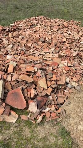 The video shows a large pile of broken terracotta tiles and some concrete fragments in an open, grassy field. The pile is extensive, covering a significant area of the ground. The terracotta pieces vary in size and shade, from reddish-orange to darker, weathered tones, and some lighter-colored, possibly concrete or cement, chunks are interspersed throughout. The surrounding ground is covered in green grass with some patches of dry, light brown soil visible. The lighting suggests it is daytime with clear weather. There are no people or discernible activities in the video. The setting appears to be rural, possibly in Meore Kesalo, Georgia, given the location context. No text is visible.